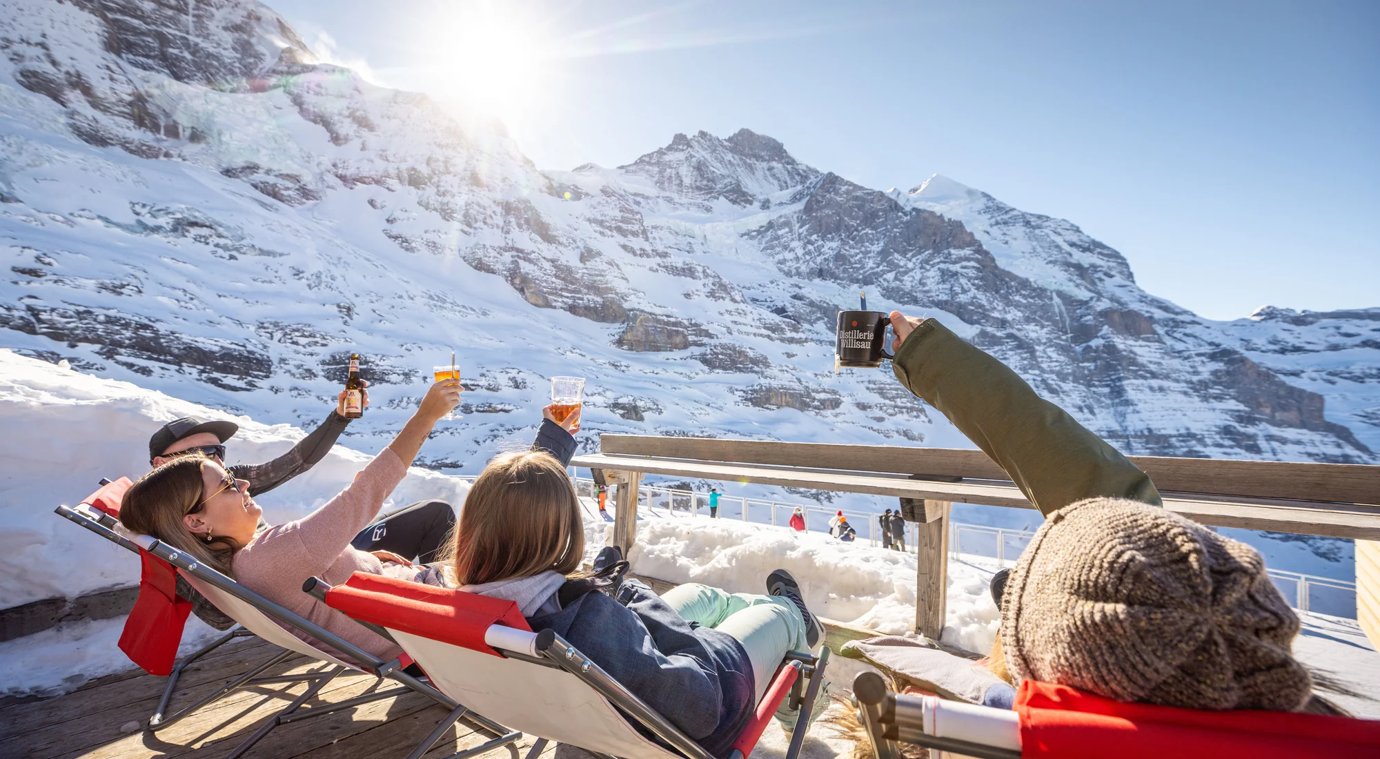Skiers relaxing in deckchairs with drinks in spring sunshine above Lauterbrunnen, snow-capped Jungfrau peaks behind