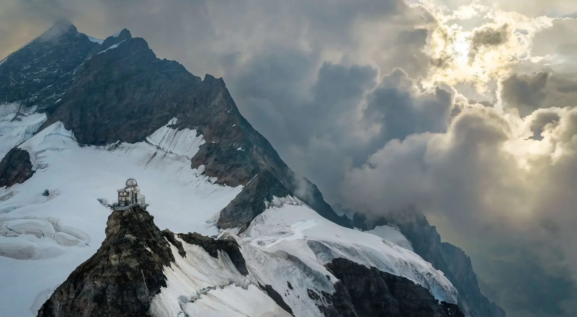 Das Observatorium Sphinx auf seinem Felssporn am Jungfraujoch, mit dichten Wolken rund um den Gipfel und Sonne, die dahinter durchbricht