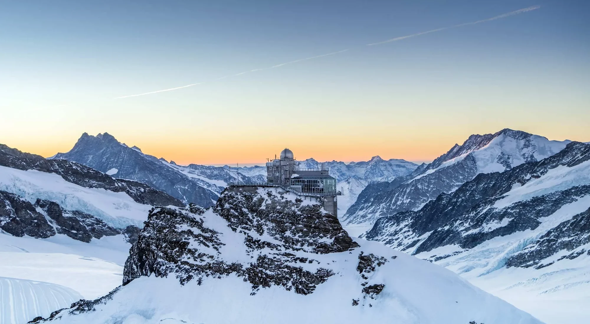El observatorio Sphinx del Jungfraujoch con las primeras luces, nieve y glaciar extendiéndose bajo un cielo despejado de amanecer anaranjado