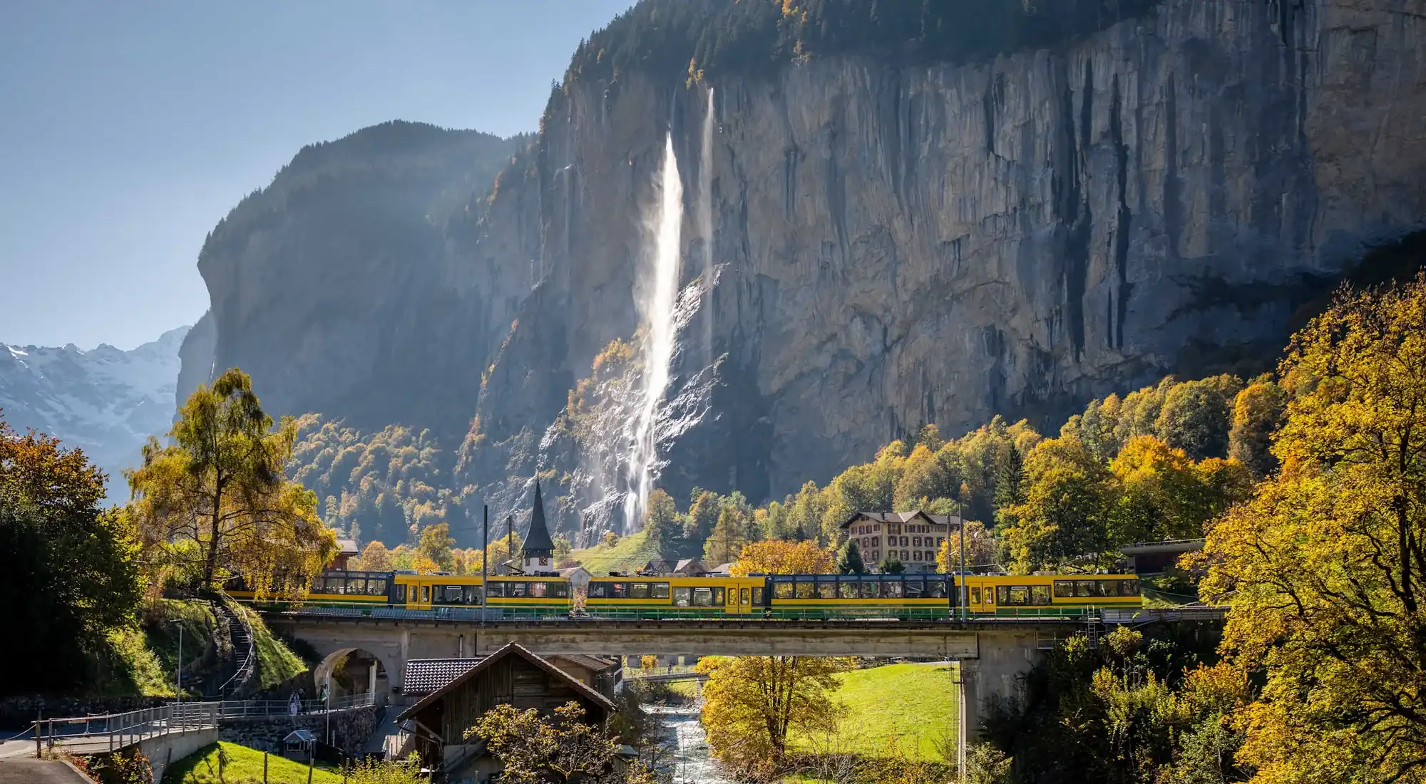 El valle de Lauterbrunnen en otoño, un tren amarillo de la Wengernalpbahn cruzando el puente bajo las cataratas del Staubbach con el campanario y los árboles dorados