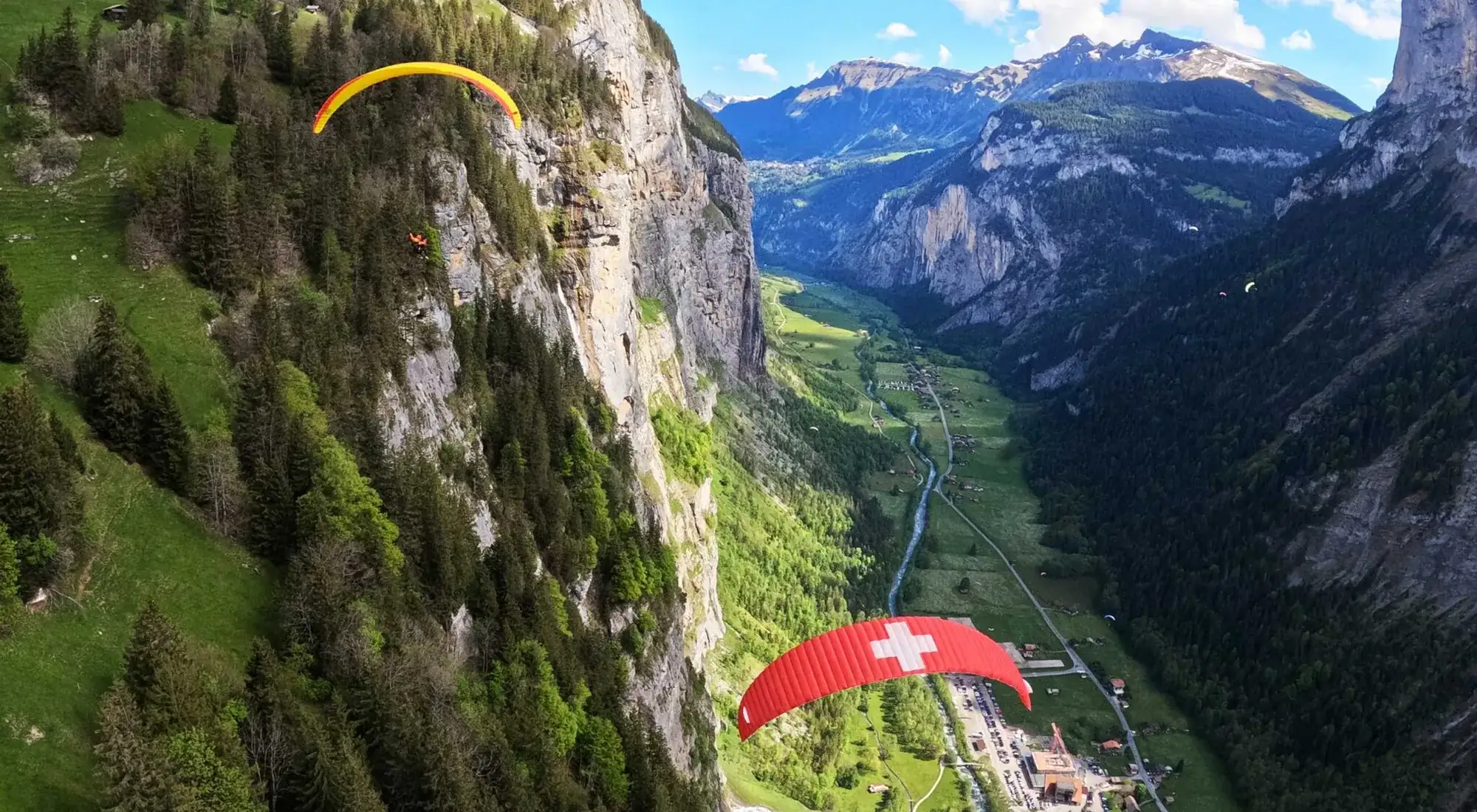 Zwei Tandem-Gleitschirme über dem Lauterbrunnental, ein gelber Schirm nah an der Felswand unter Mürren und ein roter Schirm mit Schweizerkreuz weiter unten, Talboden und Fluss weit darunter