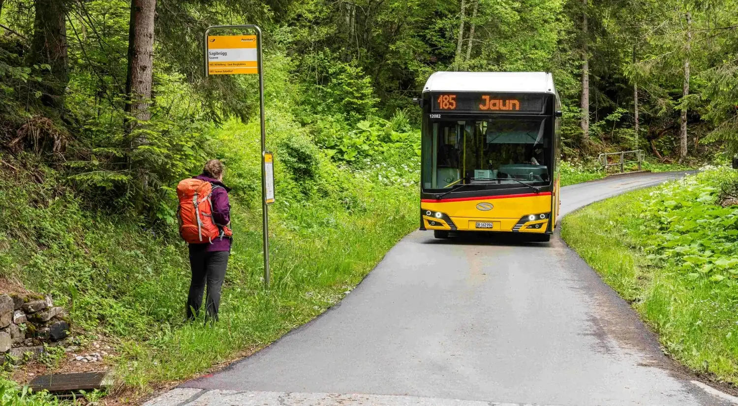 Ein gelbes Postauto, das an einer Haltestelle am Strassenrand einfährt, wo ein Wanderer mit Rucksack wartet, Wald auf beiden Seiten